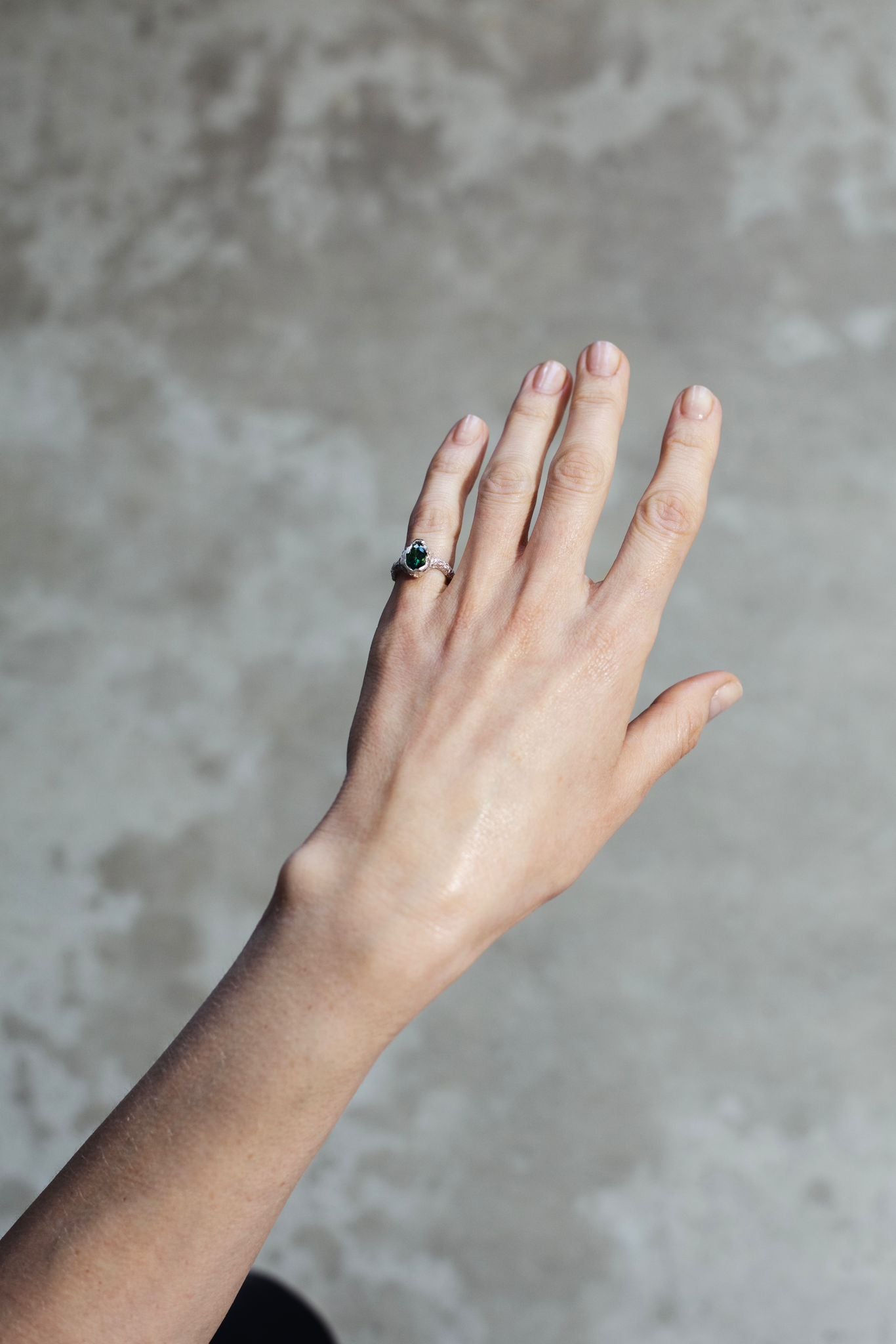 The image depicts a human hand raised against a grey background. The hand is positioned with the palm facing towards the viewer and fingers extended upwards. The small finger has a bulky silver ring with an deep green coloured stone.